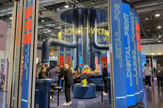Trade fair stand with a blue circular structure and suspended pipes; visitors among tall banners with text about digital printing and retail.